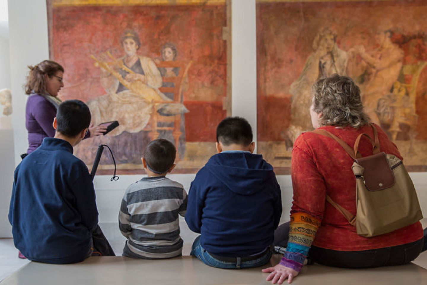 Four people, two with guide dogs, walk from right to left in front of a large tapestry. They wear headphones around their necks and carry bags.